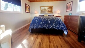 Un dormitorio con techo de madera, una cama con colcha de rayas azules y blancas, dos lámparas de noche, un aparador y cuadros en las paredes.