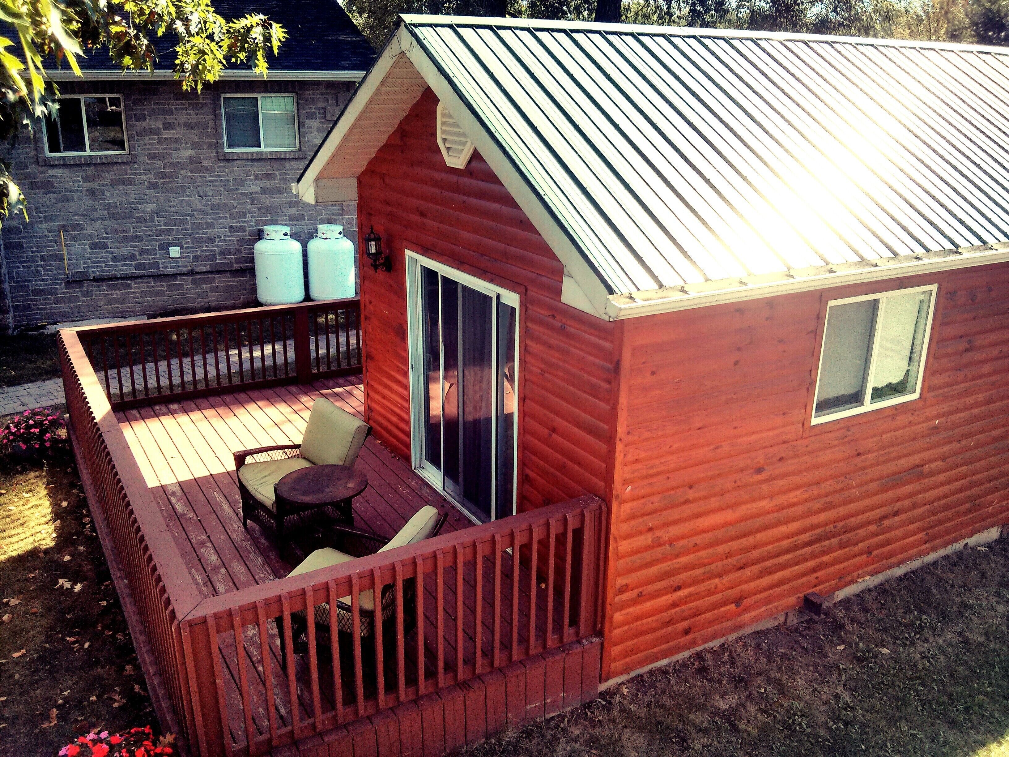 Una pequeña cabaña de madera con terraza, puerta corrediza de vidrio y dos tanques de propano grandes afuera.