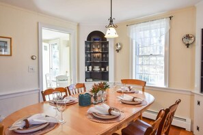 Une salle à manger avec une table en bois dressée pour six personnes, entourée de chaises. Un lustre est suspendu au plafond, et une fenêtre avec des rideaux légers est visible.