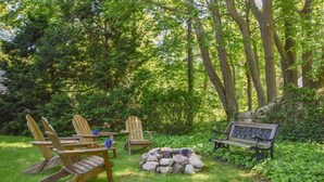 A garden with wooden chairs and a bench, a fire pit, and dense greenery.
