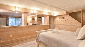 Quarto com piso de madeira, cama com roupa de cama branca, escrivaninha de madeira e janela com cortinas.