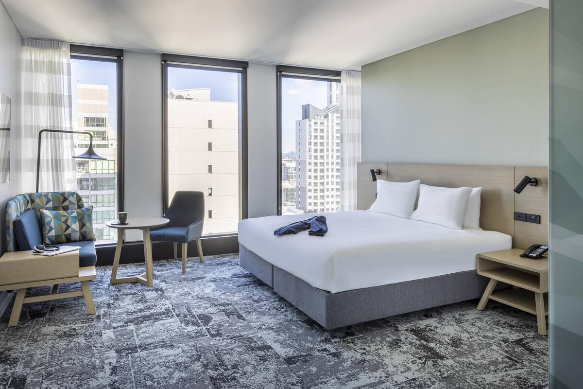 A modern hotel room with a large bed, a desk, a chair, and a view of the city through large windows.