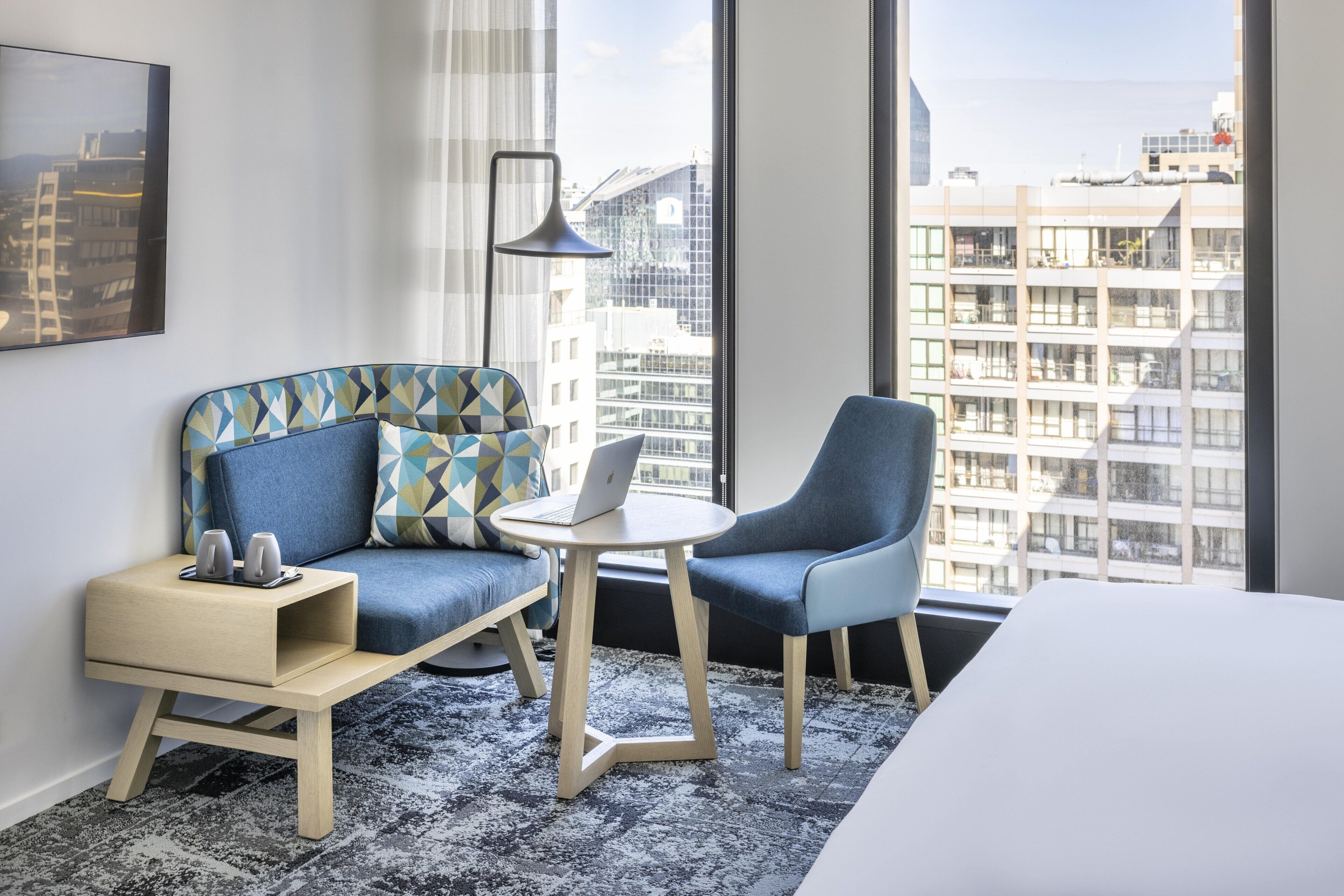 A modern hotel room with a sofa, a chair, a small table, and a large window offering a city view.