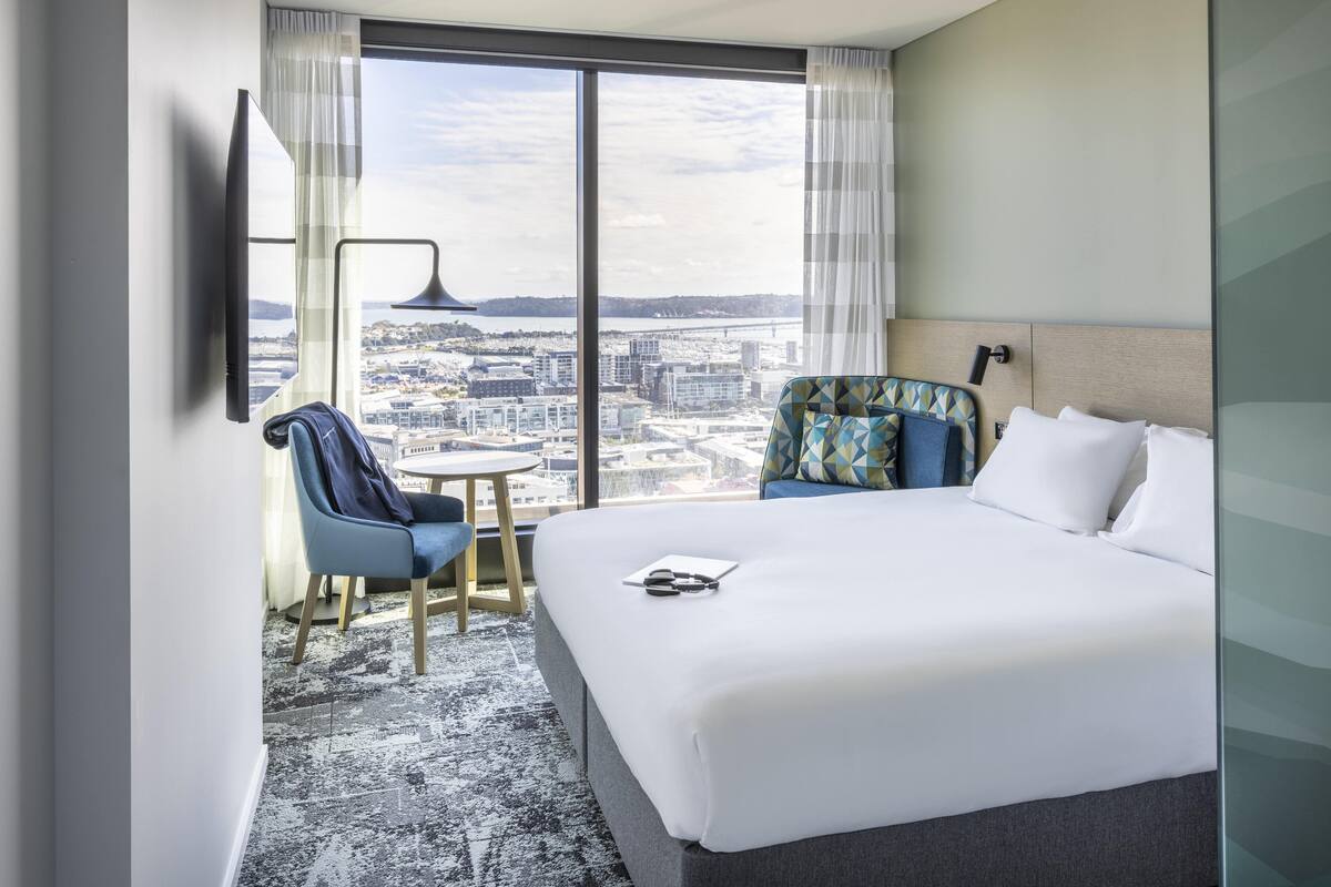 A hotel room with a large bed, a chair, a small table, a TV mounted on the wall, and a view of the city through large windows.