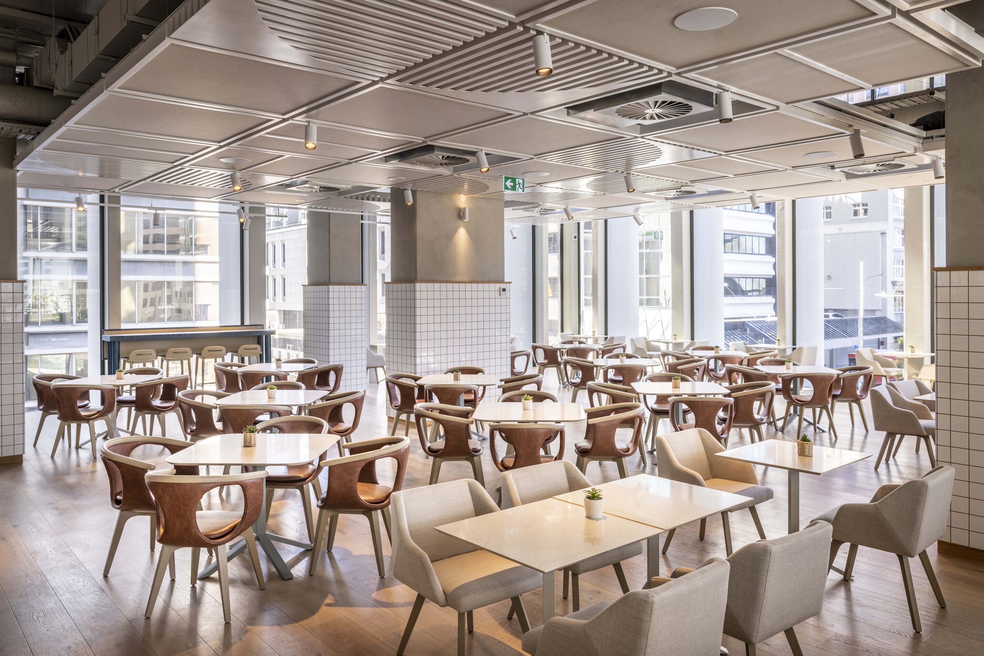 A spacious dining area with multiple tables and chairs, large windows, and a modern ceiling design.
