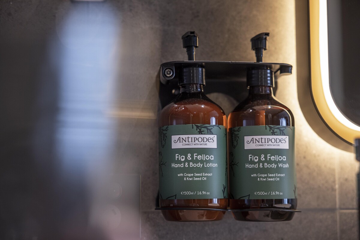 Deux flacons de lotion pour les mains et le corps Antipodes, parfumée à la figue et au goyavier, et un flacon de gel lavant pour les mains et le corps, présentés sur un distributeur mural.