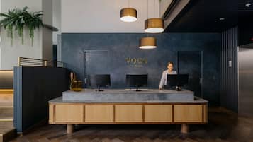 Un mostrador de recepción de diseño moderno, con una pared oscura que muestra el nombre "VOCO" y dos monitores. Debajo del mostrador hay armarios de madera y una planta a la izquierda.