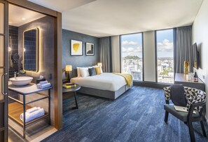 A modern hotel room with a large bed, a sitting area, a bathroom with a sink and mirror, and a city view. - voco Auckland City Centre by IHG (Auckland)