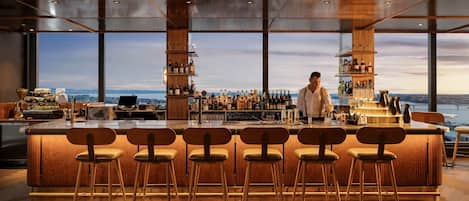 Eine Bar mit einer hohen Theke, mehreren Hockern und einem Barkeeper. Die Bar hat eine Holzinnenausstattung und große Fenster mit Blick auf die Stadt.
