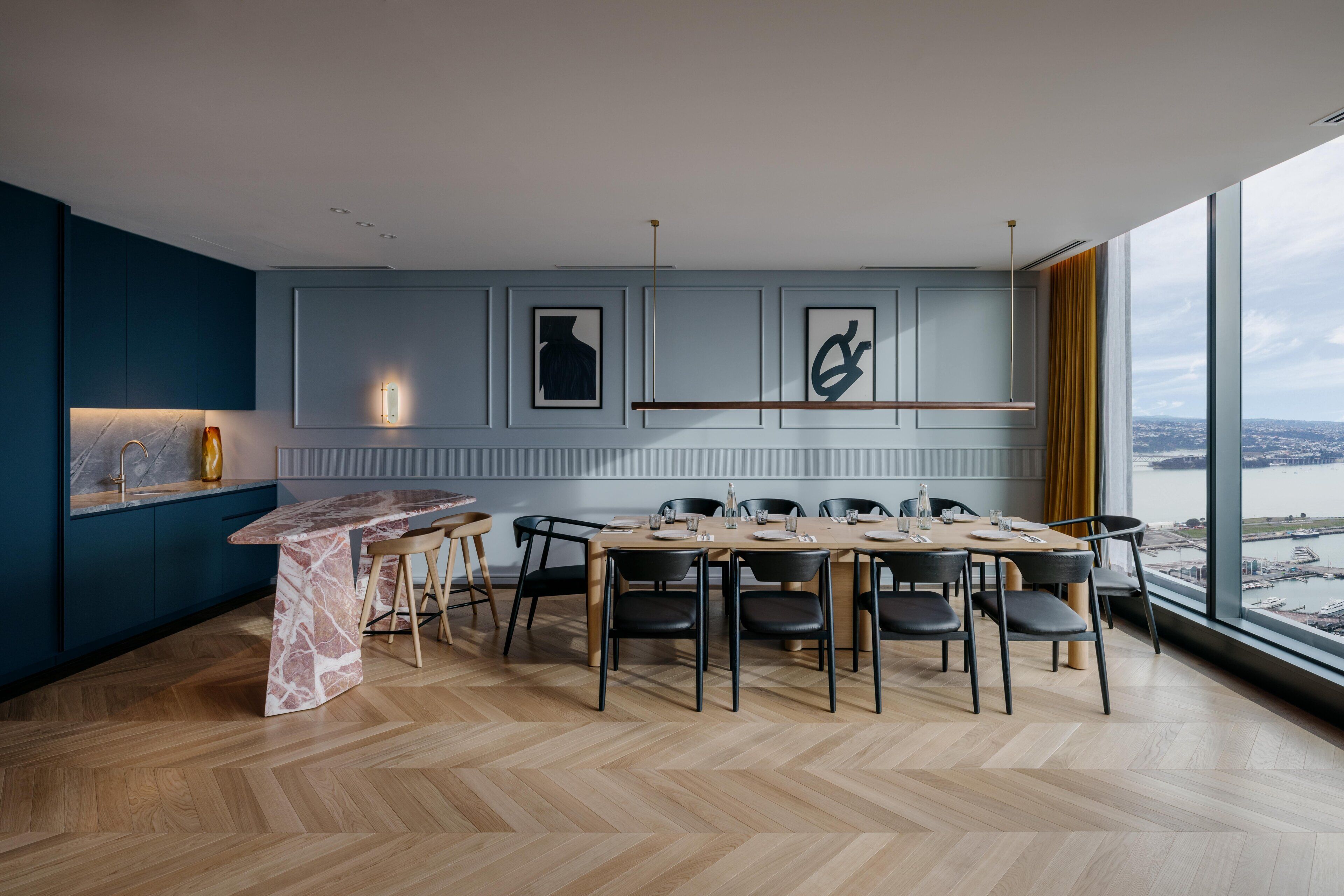 A modern dining area with a marble island, wooden chairs, and a view of a body of water through large windows.