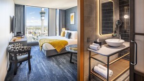 A modern hotel room with a bed, a desk, and a view of a cityscape including a tower. - voco Auckland City Centre by IHG (Auckland)