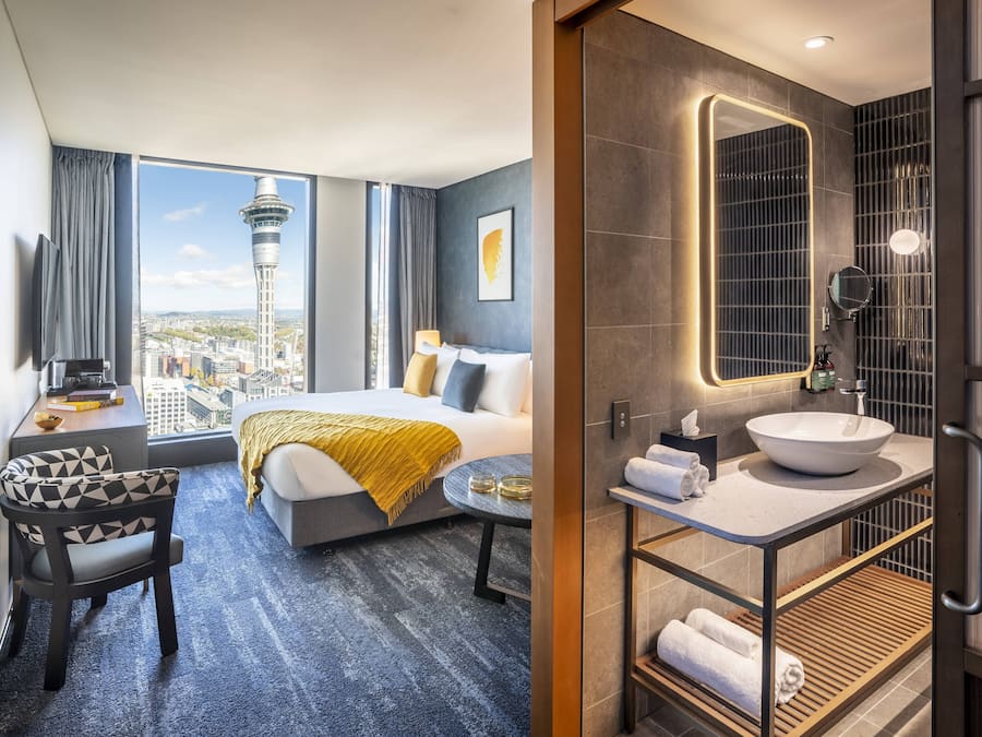 A modern hotel room with a bed, a desk, and a view of a cityscape including a tower.