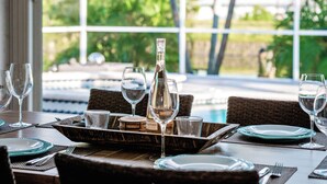 Mesa de comedor preparada con platos, vasos y una botella de vino, con vista a una piscina y follaje exterior.