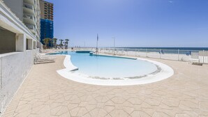 A swimming pool with a curved design, surrounded by a tiled deck and a white fence. - Ocean House 1304 | Heated, Zero Entry Pool | Oceanfront Balcony | My Beach Getaways (Gulf Shores)