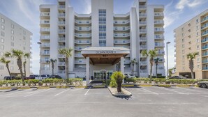 A multi-story hotel with a central entrance, balconies, and a parking lot with palm trees. - Ocean House 1304 | Heated, Zero Entry Pool | Oceanfront Balcony | My Beach Getaways (Gulf Shores)
