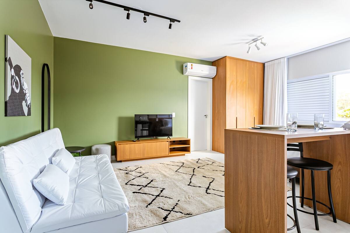 Una sala de estar moderna con un sofá blanco, un televisor sobre un mueble de madera y una zona de cocina con barra.