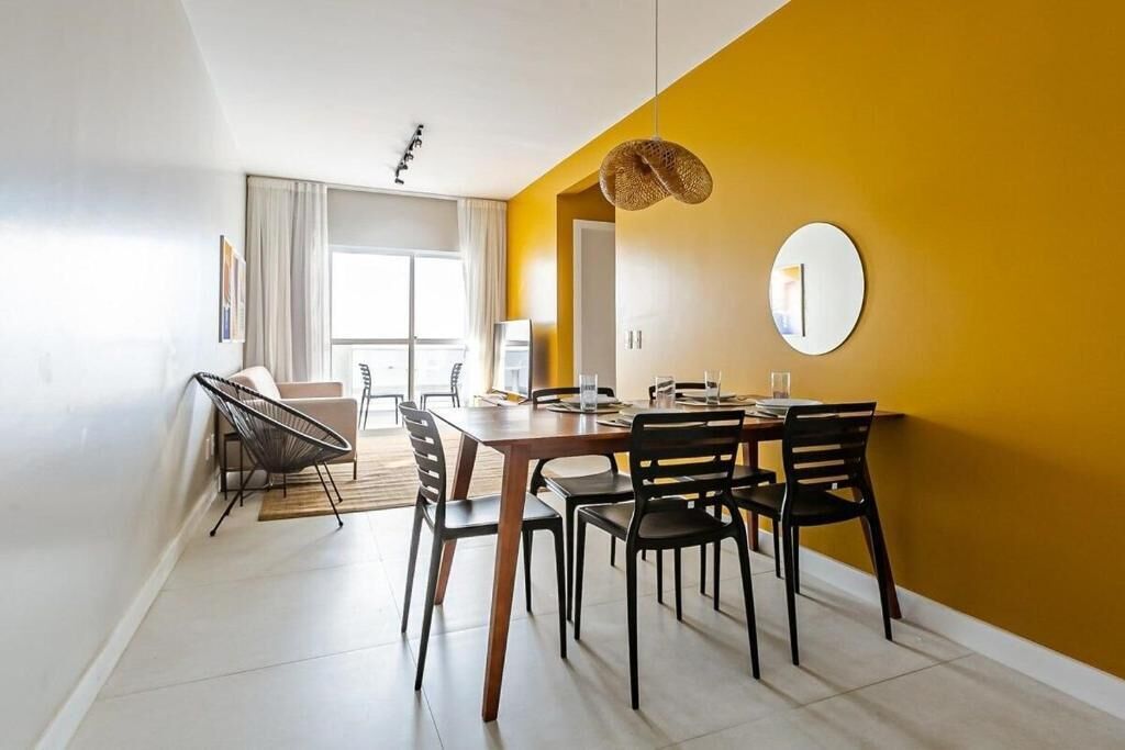A modern dining area with a wooden table and black chairs, a round mirror on the wall, and a yellow accent wall.