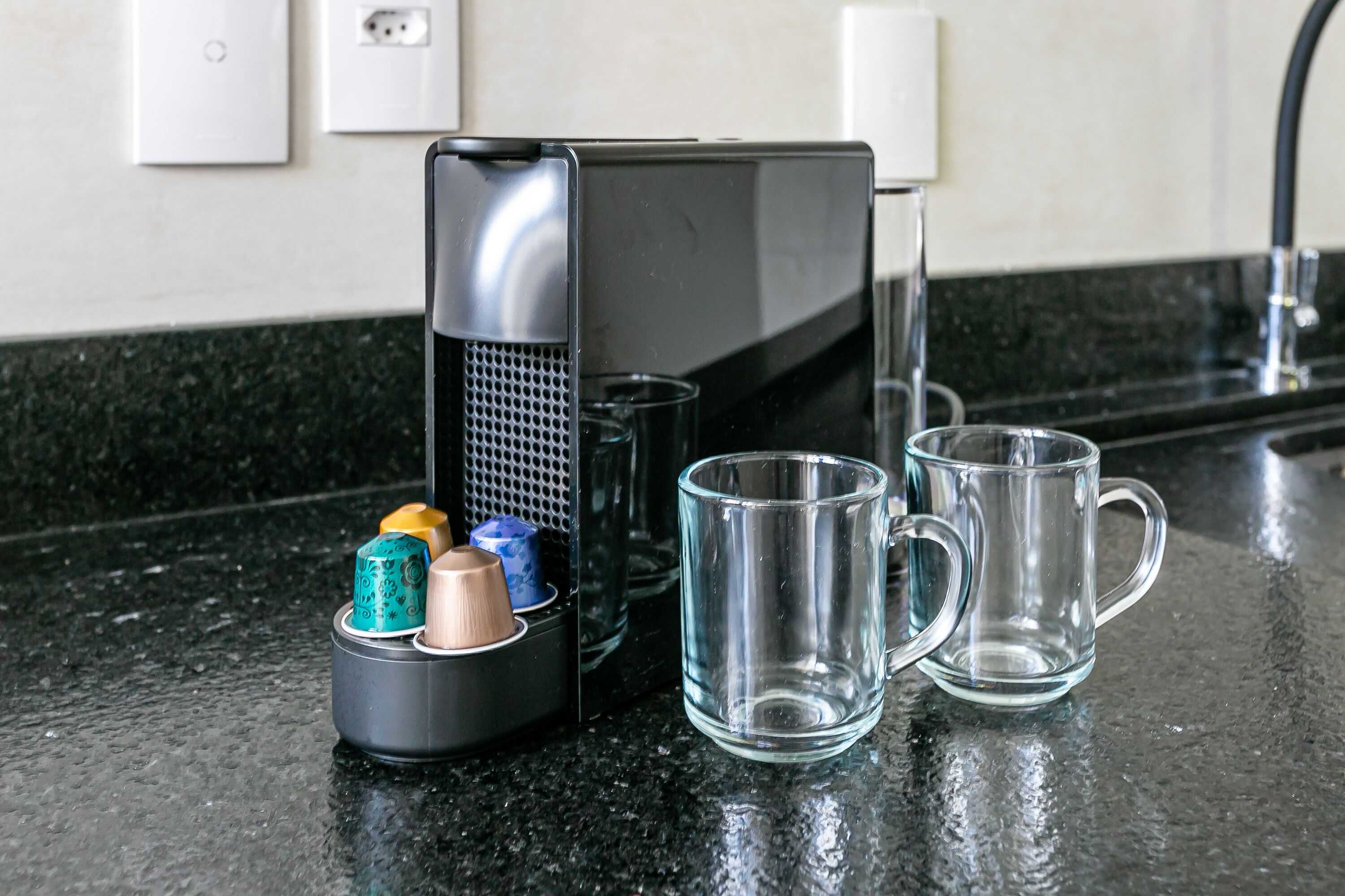 A kitchen counter with a coffee maker, a glass container with utensils, and two empty glass mugs.