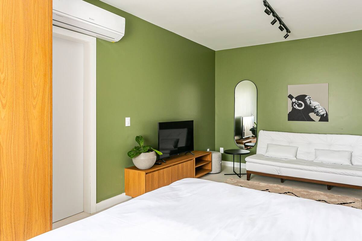 Un dormitorio moderno con cama, televisor sobre un mueble de madera, sofá blanco y pared verde.