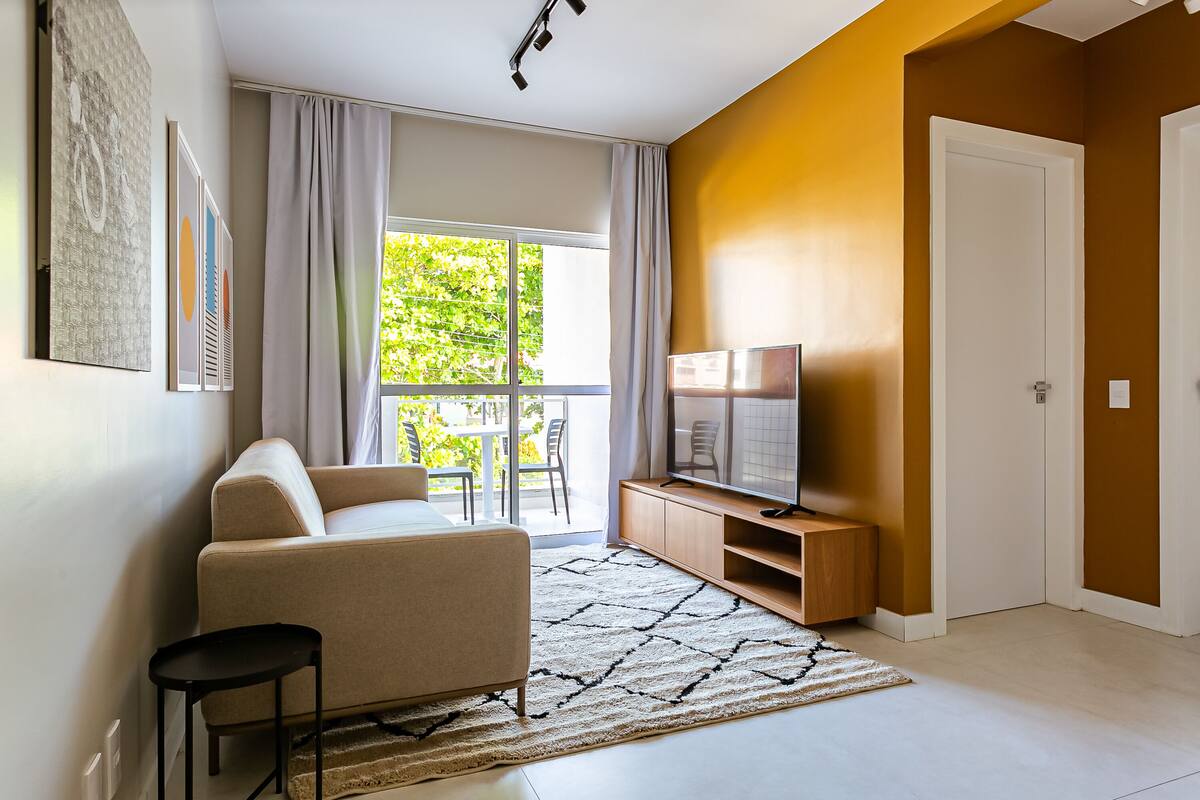 Una sala de estar moderna con un sofá, un televisor sobre un mueble de madera, una mesita y un ventanal con cortinas.