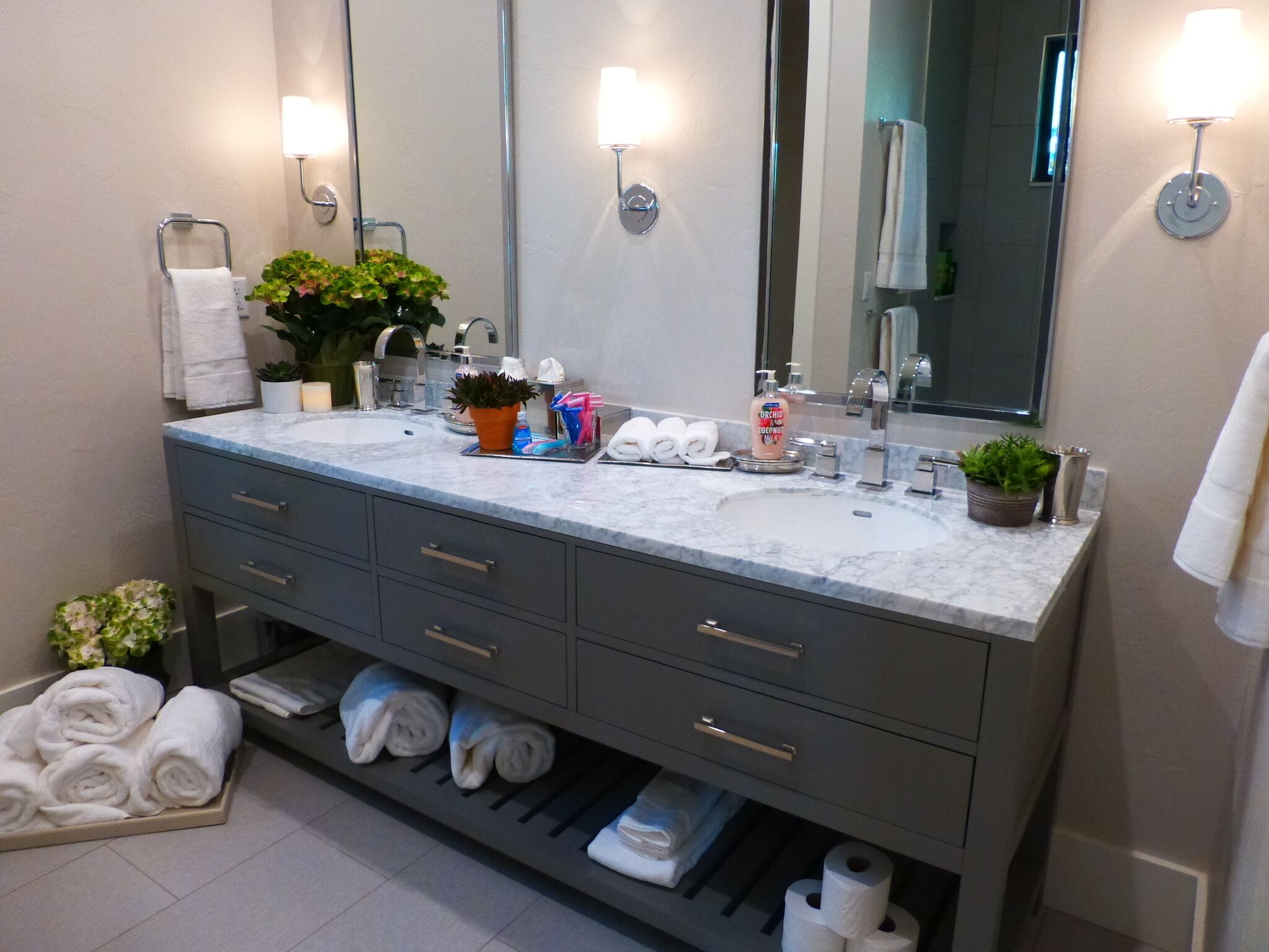 A modern bathroom with a marble sink, two mirrors, a towel rack, a plant, and neatly folded towels.