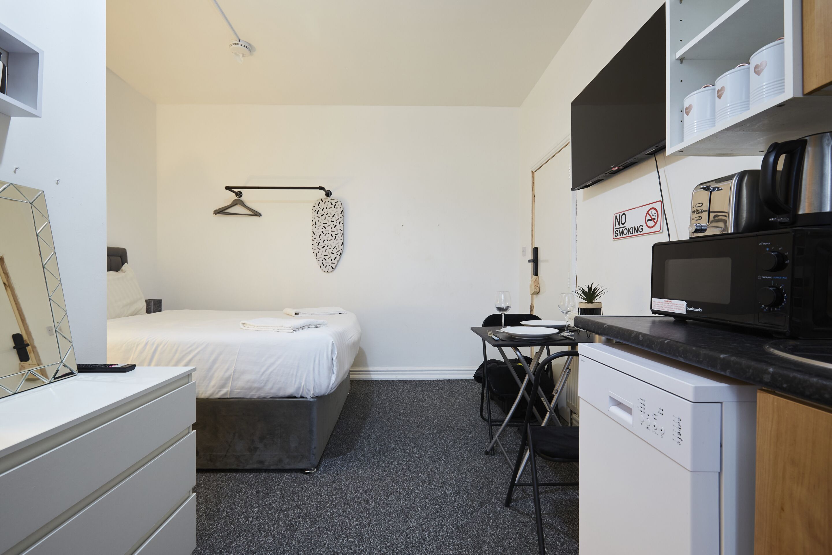 Habitación de hotel compacta con cama, una pequeña mesa con sillas, microondas, cafetera, planta colgante y un cartel que indica 'No Fumar'.