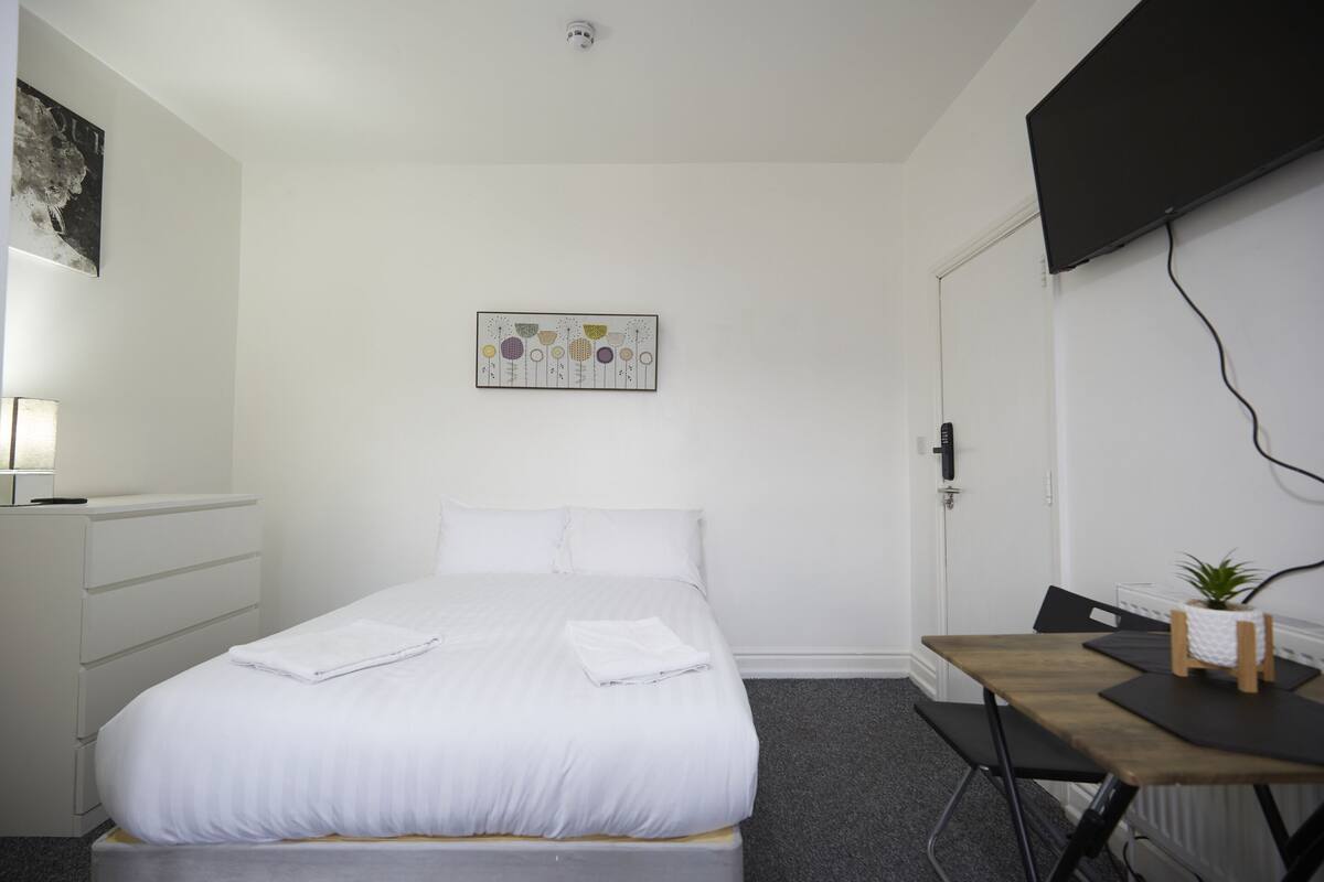 Un dormitorio con cama, escritorio, silla, una planta pequeña y un cuadro en la pared.