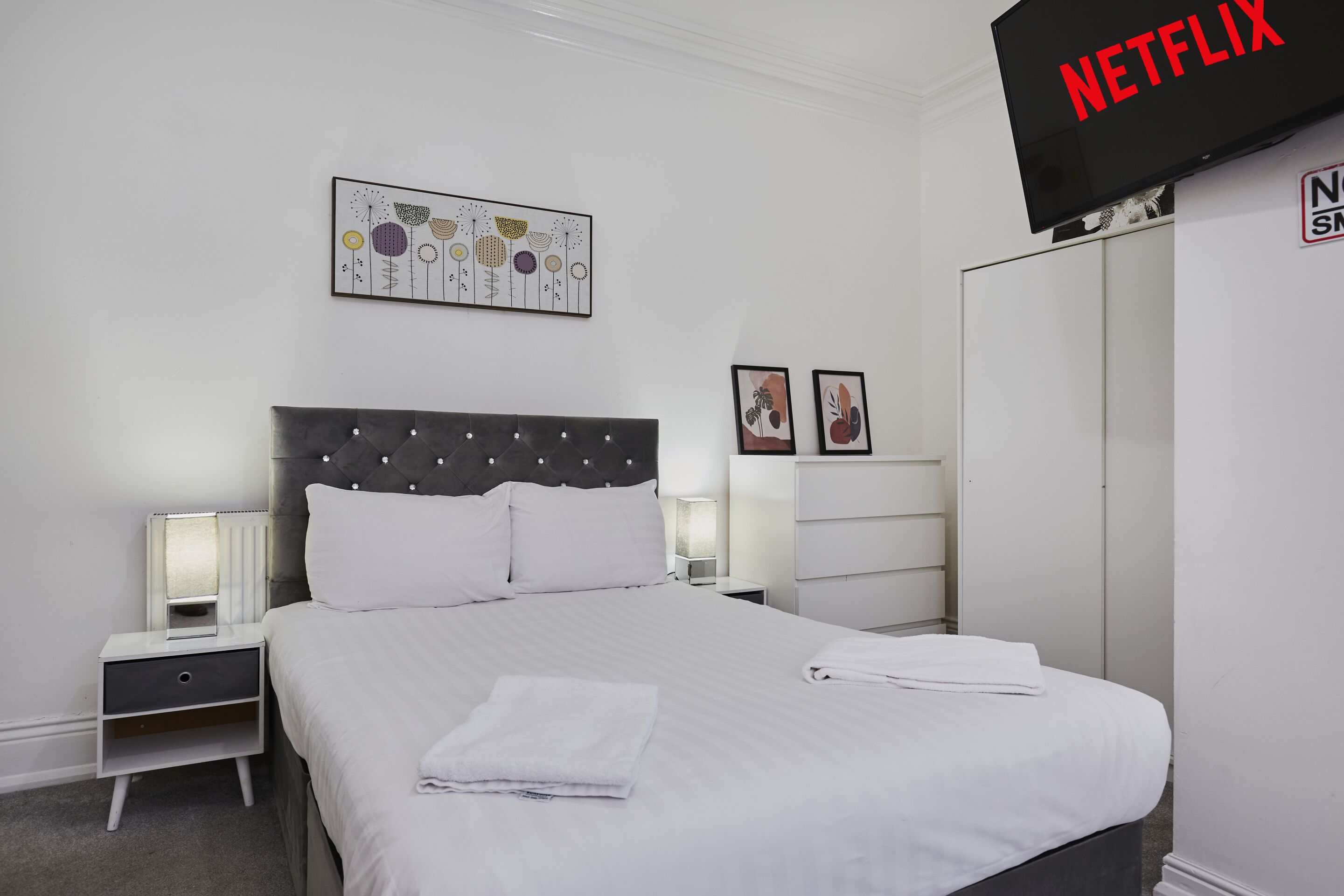A hotel room with a bed, bedside tables, a wardrobe, a TV, and artwork on the wall.