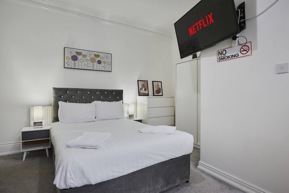 Una habitación de hotel con una cama, un televisor montado en la pared, un cartel de prohibido fumar y una obra de arte enmarcada sobre la cama.