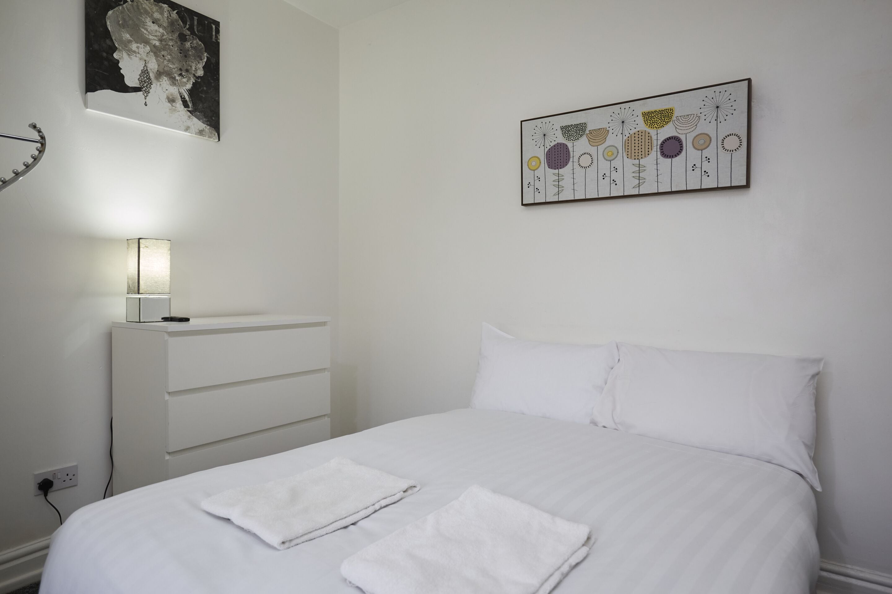 Een net opgemaakt bed met witte lakens en twee gevouwen handdoeken, een nachtkastje met een lamp, en een kunstwerk aan de muur boven het bed.