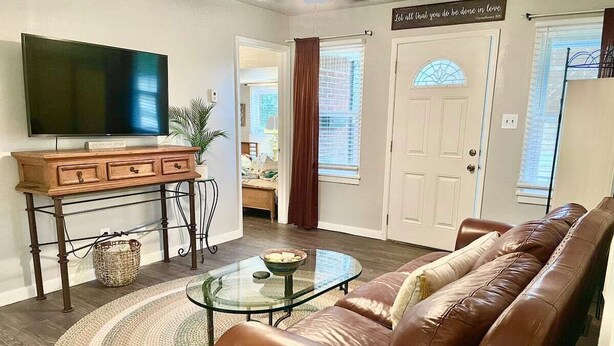 A living room with a brown leather sofa, a glass coffee table, a wooden TV stand, and a large flat-screen TV. - 3-bedroom vacation home 12 minutes from Paycom Center (Midwest City)