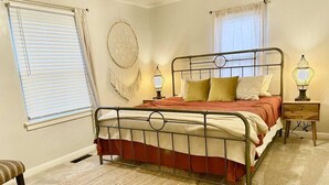 A bedroom with a metal bed frame, a red and white bedspread, two bedside lamps, a round wall hanging, and a window with blinds. - 3-bedroom vacation home 12 minutes from Paycom Center (Midwest City)