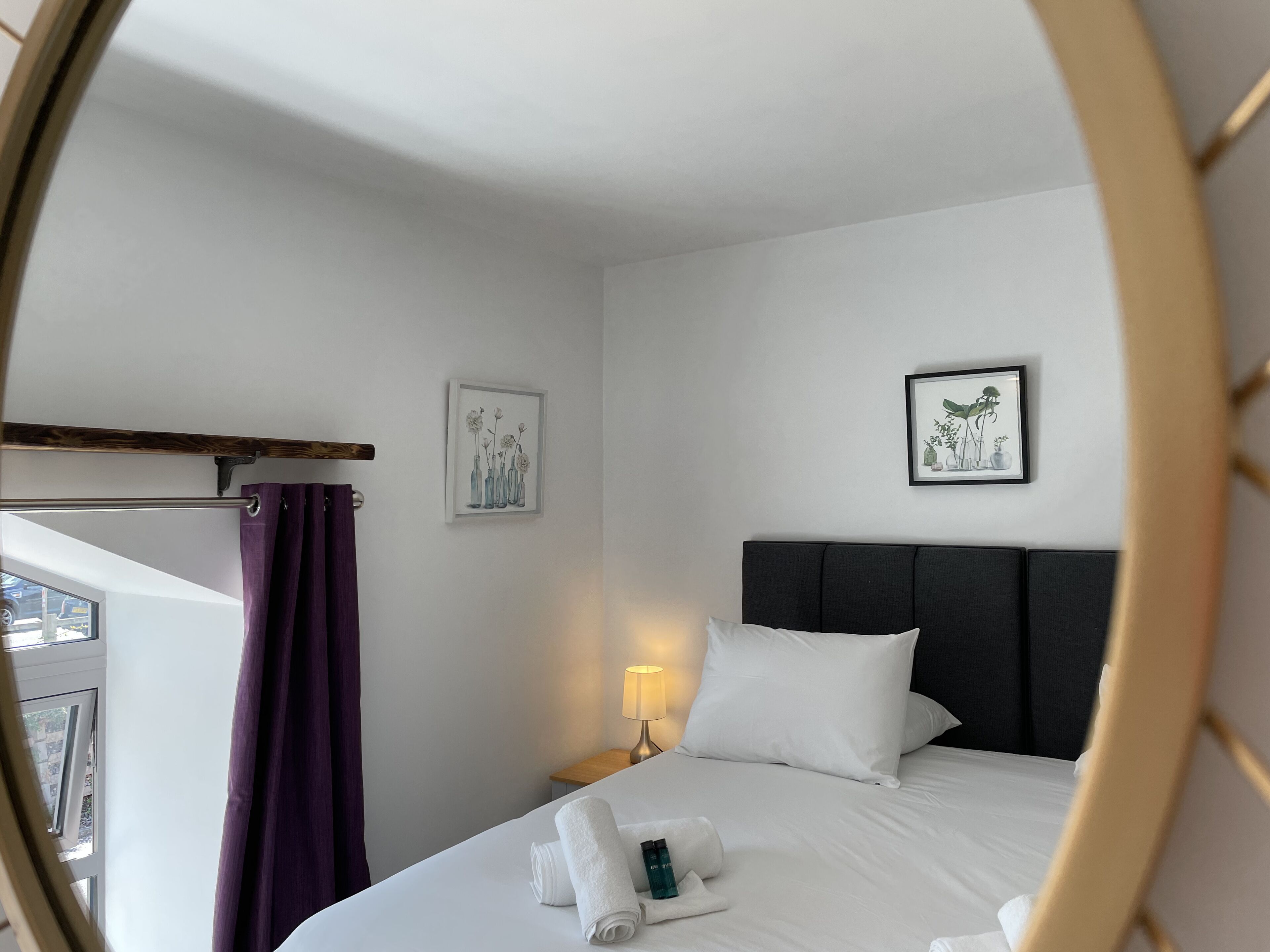 Une chambre d’hôtel avec un lit, une table de chevet avec une lampe, une fenêtre avec des rideaux violets et deux tableaux encadrés accrochés au mur.