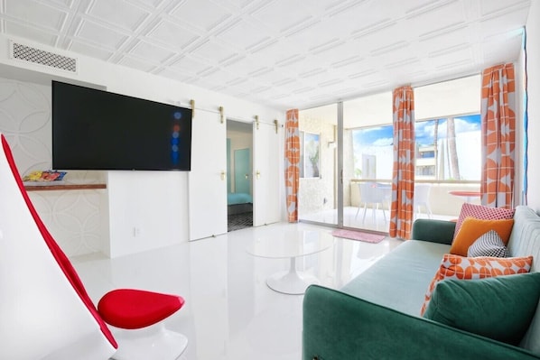 A modern living room with a flat-screen TV, a red chair, a green sofa, and a balcony with orange curtains.