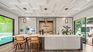 A modern kitchen with a central island, wooden chairs, pendant lights, and a view of a swimming pool.