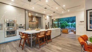 A modern kitchen with built-in appliances, a dining area with wooden chairs, and a view of greenery through large windows.