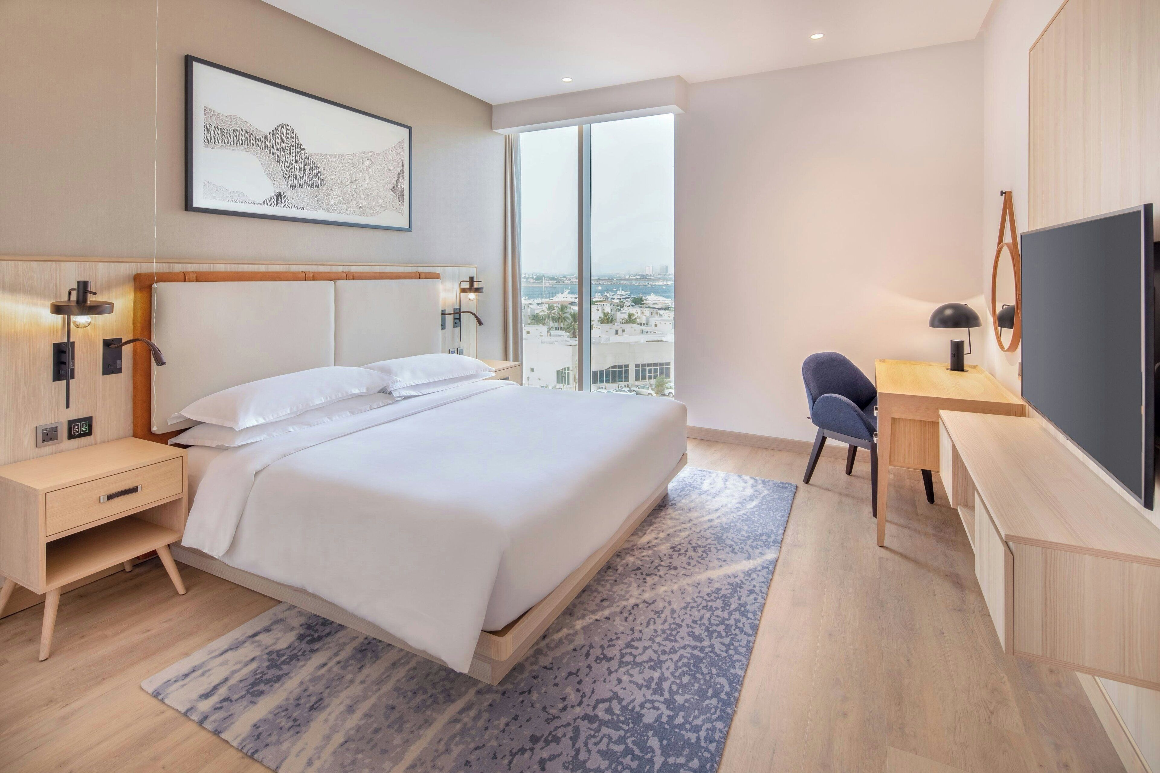 A modern hotel room with a large bed, a desk, a TV, and a view of the city.