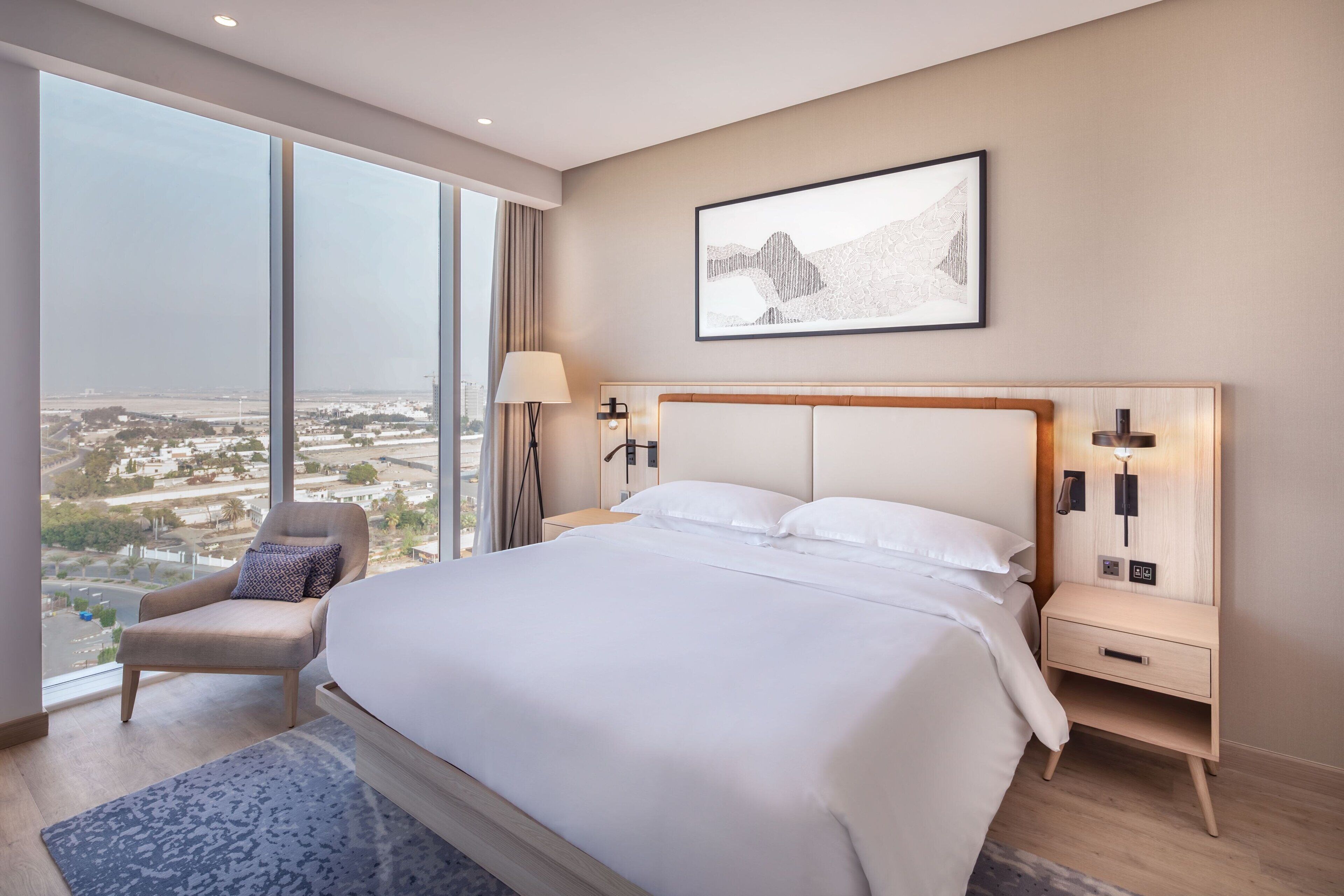 A modern hotel room with a large bed, a chair, a nightstand, and a view of the city.