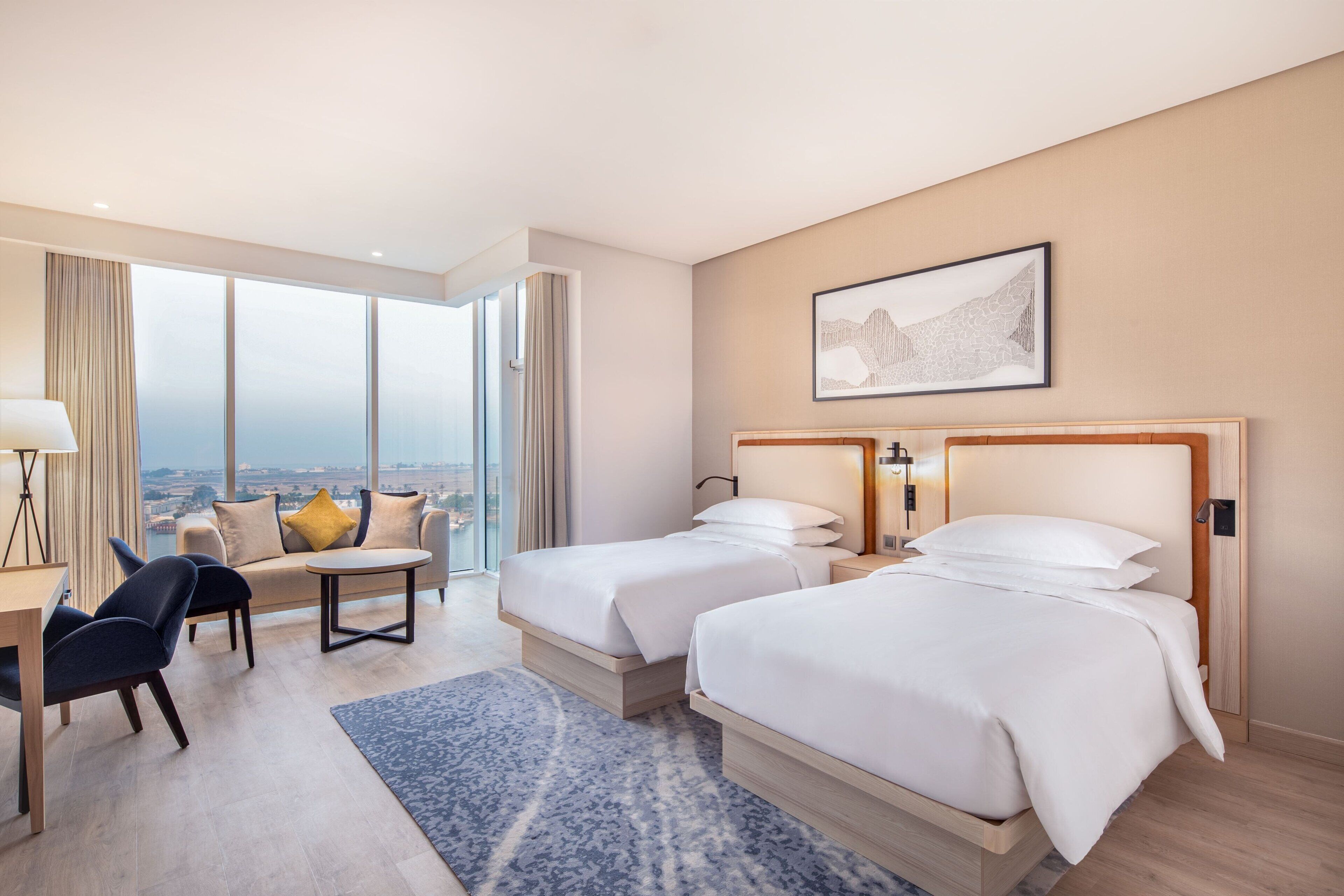 A hotel room with two beds, a sofa, a desk, and a large window offering a city view.