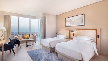 A hotel room with two beds, a sofa, a desk, and a large window offering a city view.