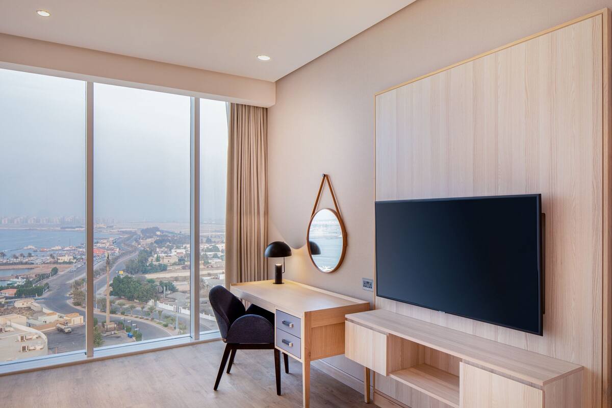 A modern hotel room with a large window offering a city view, a desk with a chair, a flat-screen TV, and a wooden panel wall.