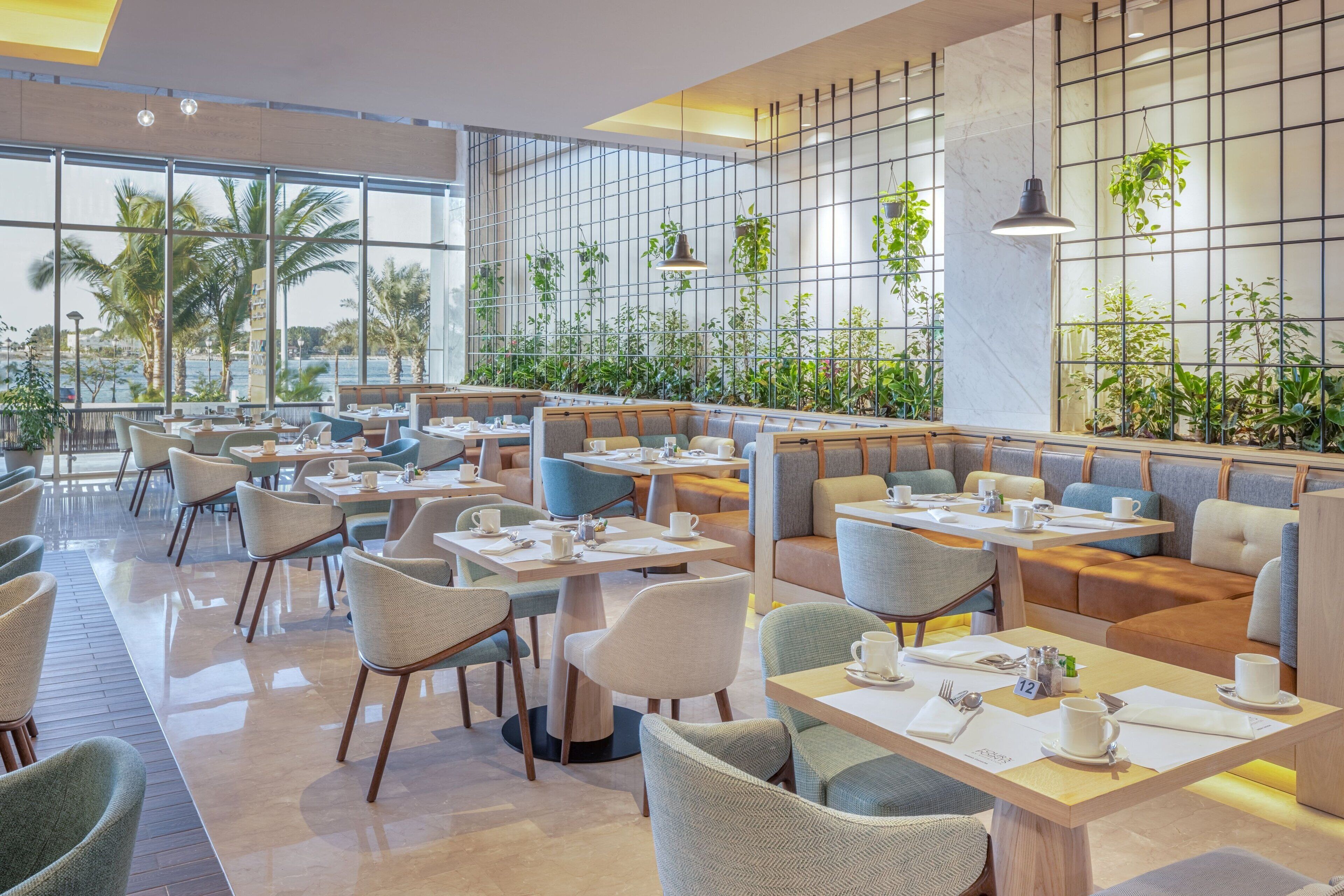 Un restaurante moderno con mesas en el interior, amplias ventanas y vistas a palmeras.