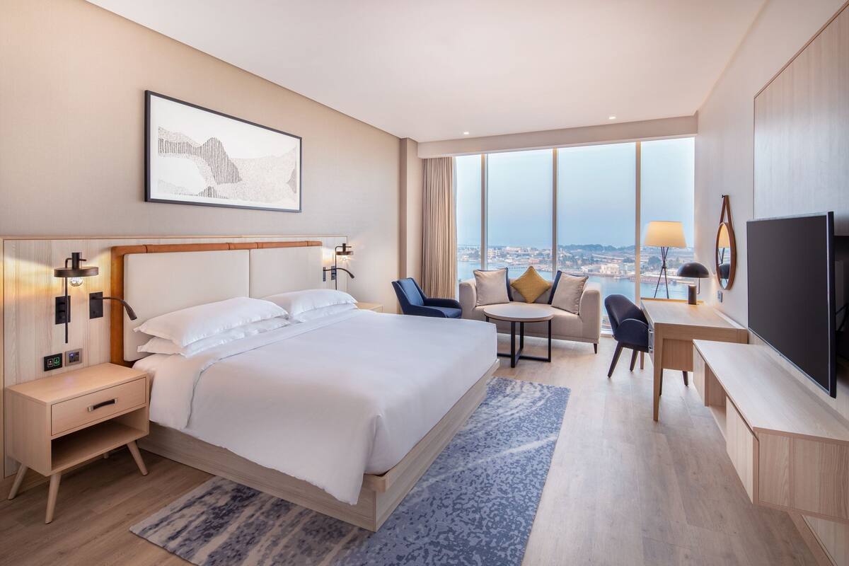 A modern hotel room with a large bed, bedside tables, a desk, a chair, a TV, and a view of the city through large windows.