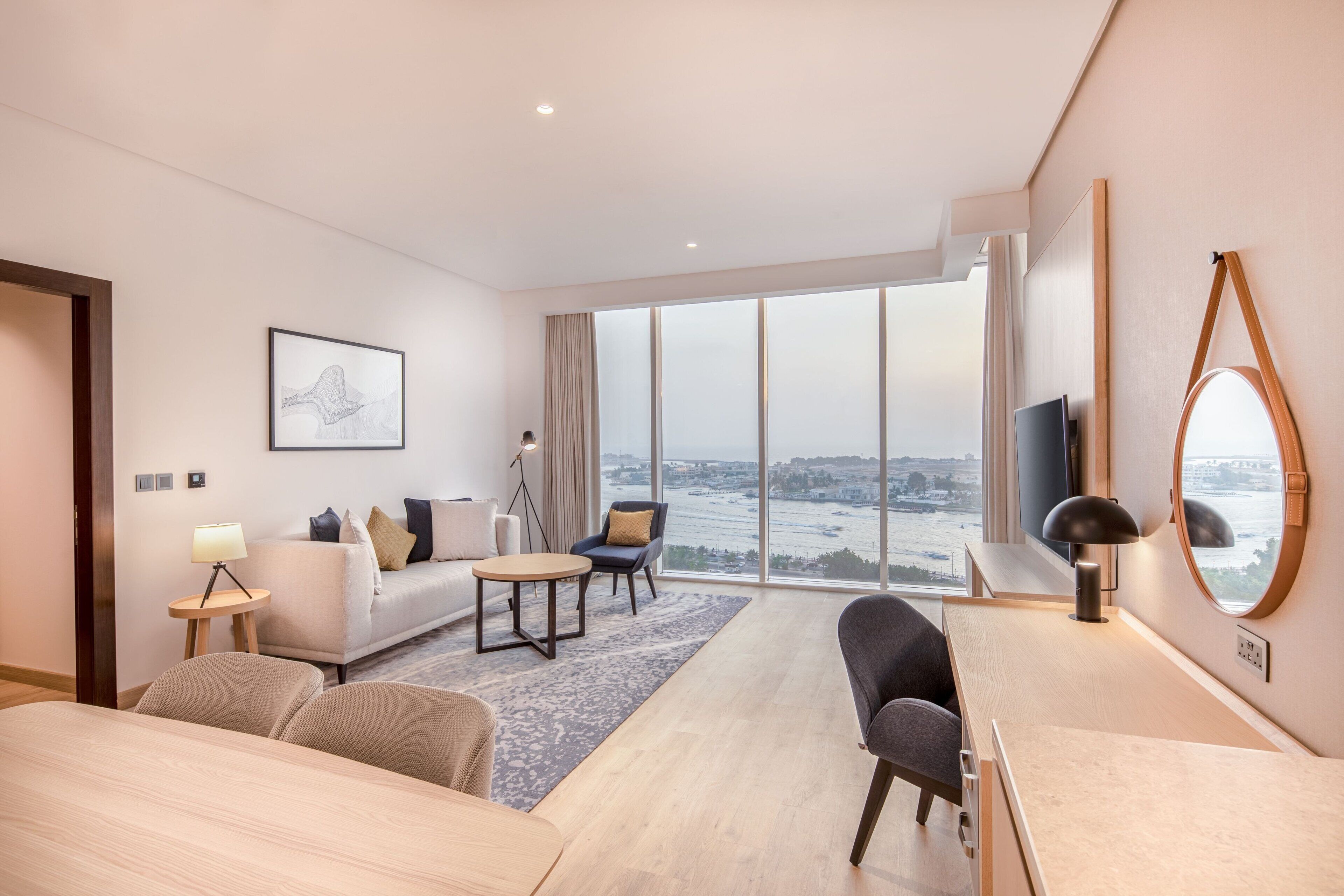 A modern hotel room with a large window offering a city view, a sofa, a dining table, and a TV.