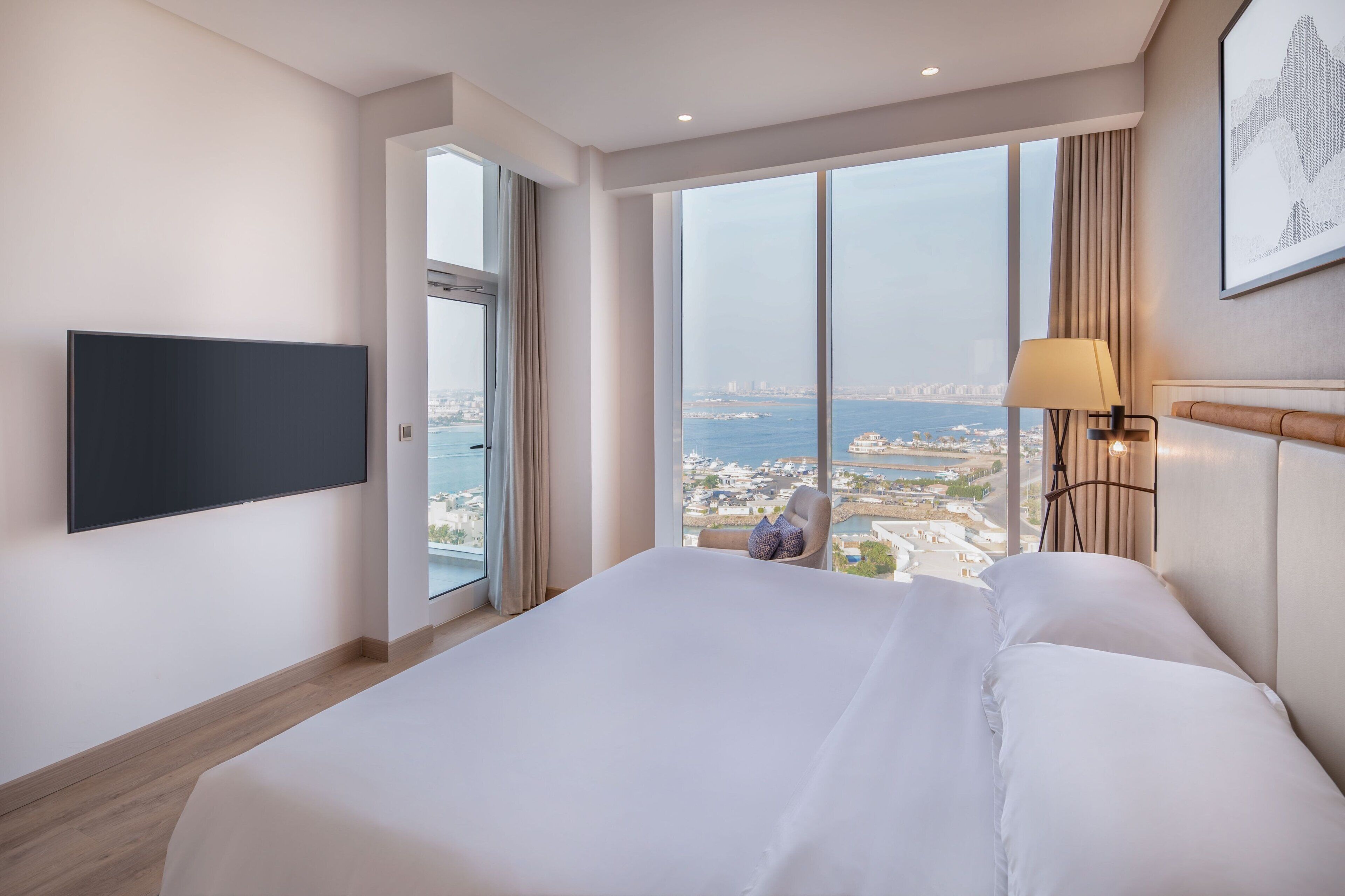 A modern hotel room with a large bed, a flat-screen TV, a city view, and a window with curtains.