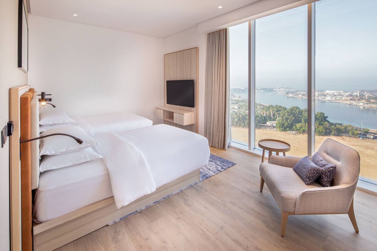 A modern hotel room with a large bed, a TV, a small table, and a view of a city and water.