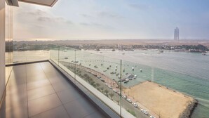 Habitación de hotel en un rascacielos con un balcón grande que da a un paseo marítimo con barcos y un paisaje urbano lejano.