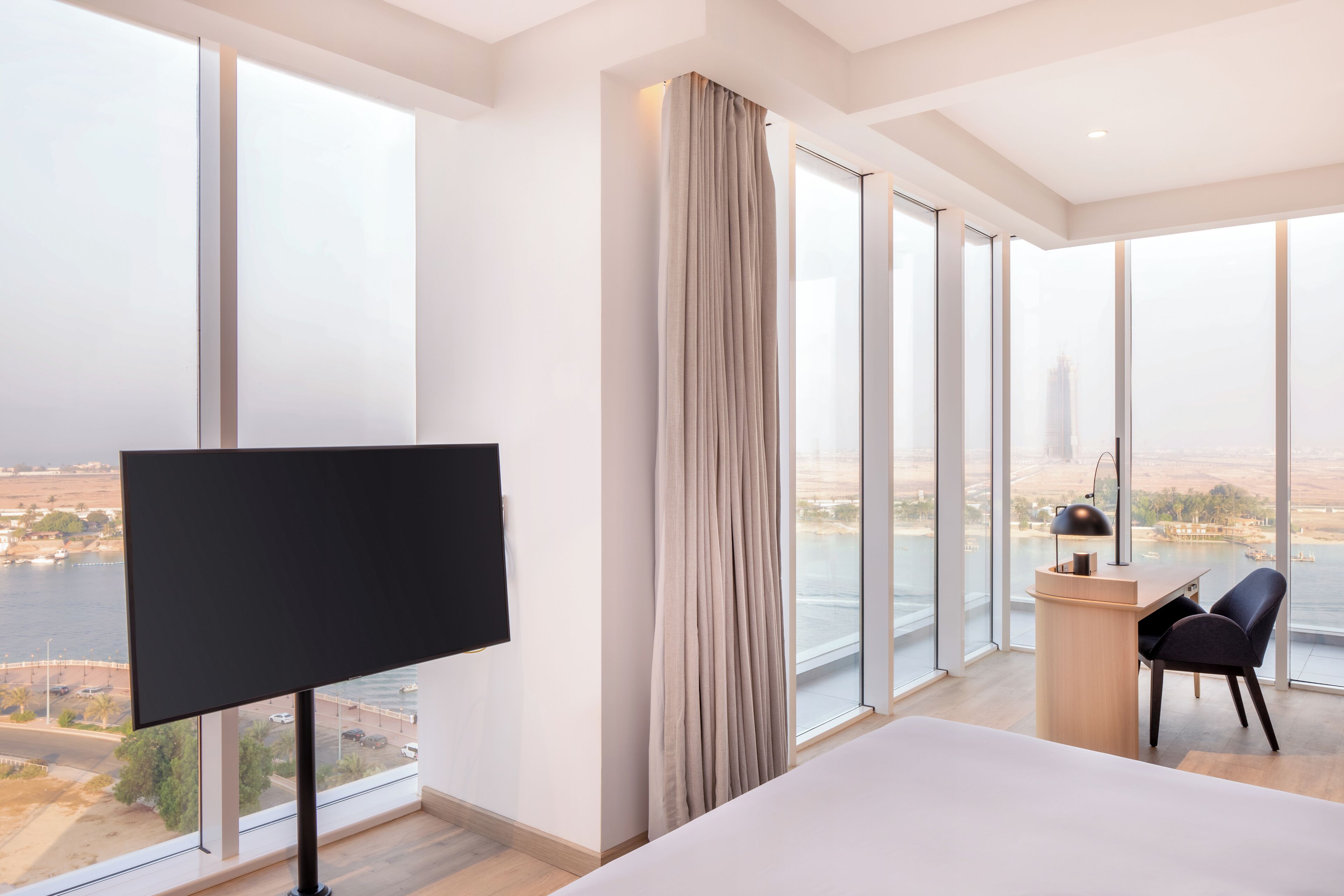 Une chambre d’hôtel moderne dotée d’un grand lit, d’un bureau avec une lampe, d’une télévision et offrant une vue sur le paysage urbain grâce à de grandes fenêtres.