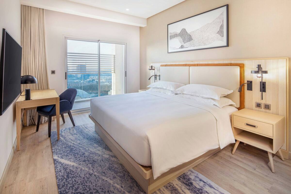 A modern hotel room with a large bed, a desk, a chair, a nightstand, and a TV.