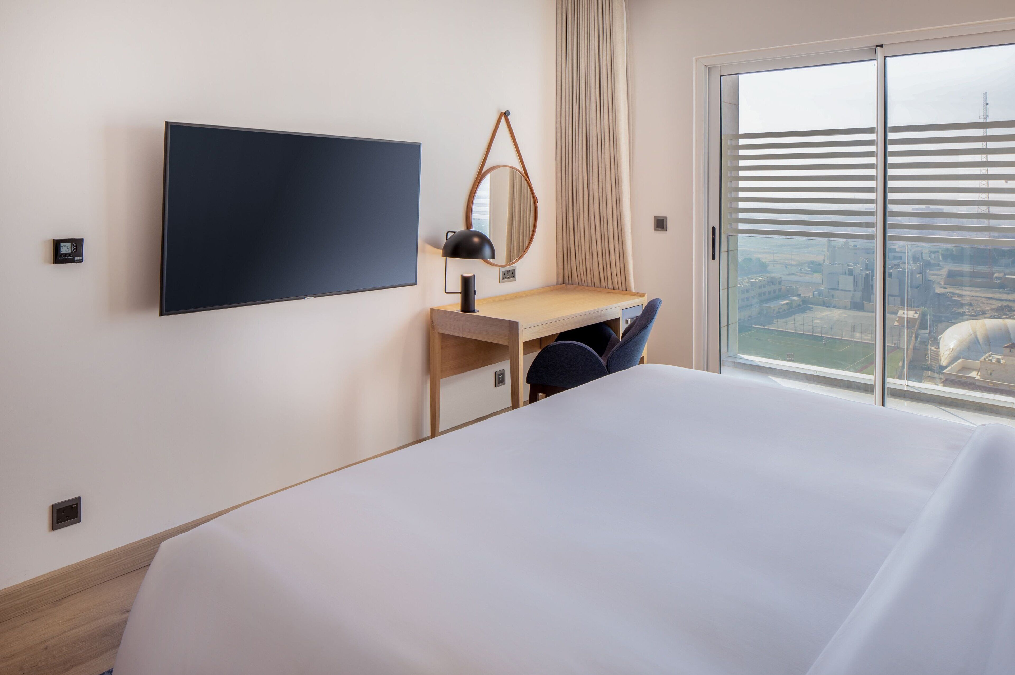 A hotel room with a large bed, a desk with a chair, a television mounted on the wall, and a window with a view of the city.